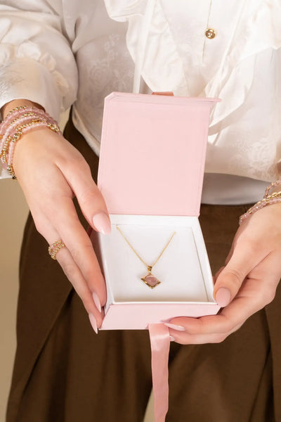 Een delicate gouden ketting met een vierkant geslepen roze edelsteen hanger, elegant tentoongesteld in een zachte roze geschenkenbox.