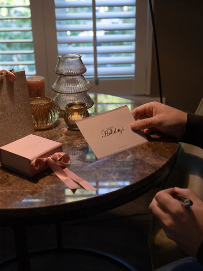 Een delicate roze groetkaart met elegante gouden cursieve letters die 'warme feestdagen' lezen en een klein logo onderaan.