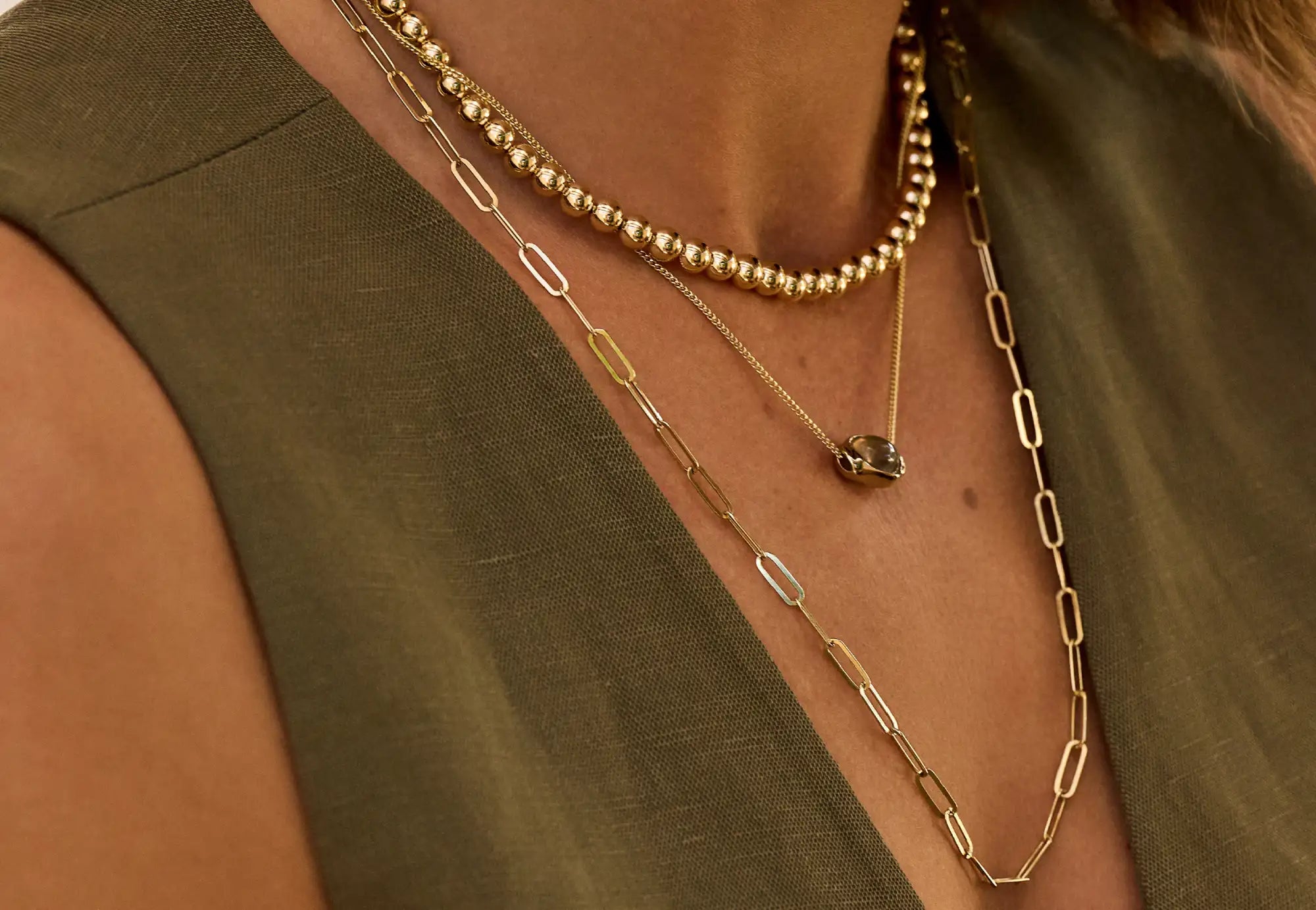 Een gelaagde set goudkleurige kettingen met paperclipkettingen, een kraalstreng en een delicate ketting met een klein hangertje.