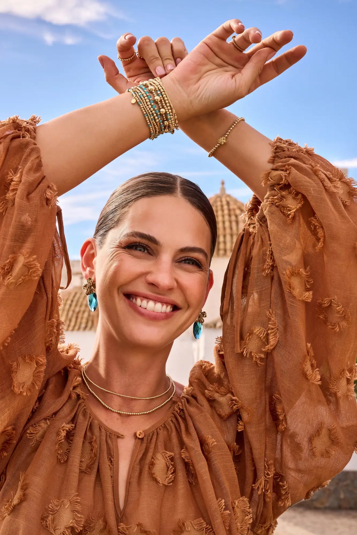 Een vrouw die een roestkleurige, gebeeldhouwde blouse draagt met geruite mouwen en gelaagde gouden kettingen.