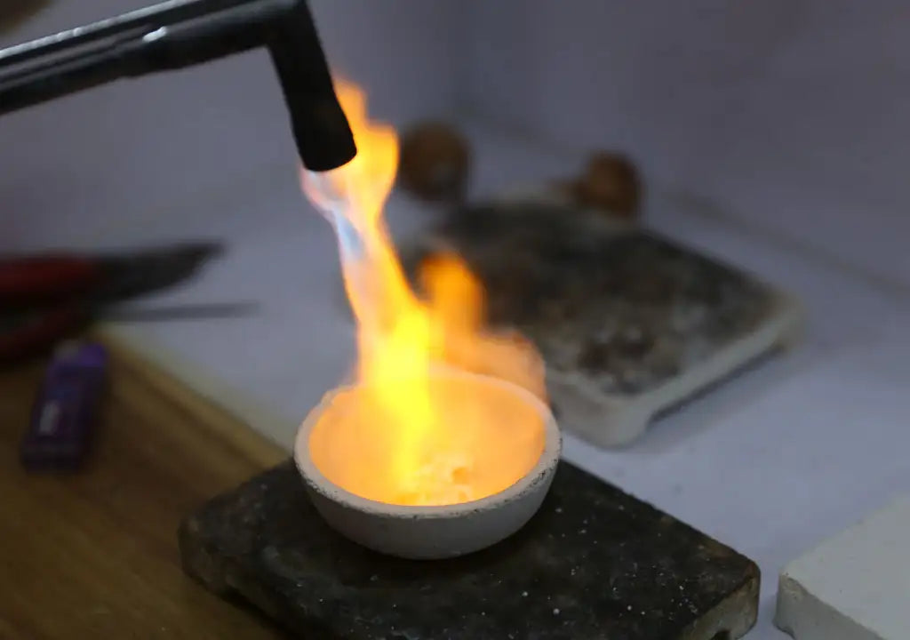 Een witte keramische smeltkroes met gesmolten metaal, verwarmd door een vlam van een toorts.